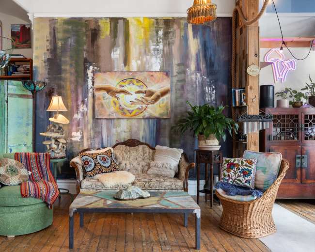 The image showcases a cozy living room with eclectic decor, featuring a patterned sofa, a wicker chair, a coffee table, and a large mural on the wall.