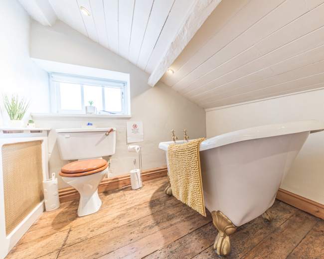 The image shows a bathroom with a freestanding bathtub on wooden flooring, a toilet, and a small window allowing natural light.