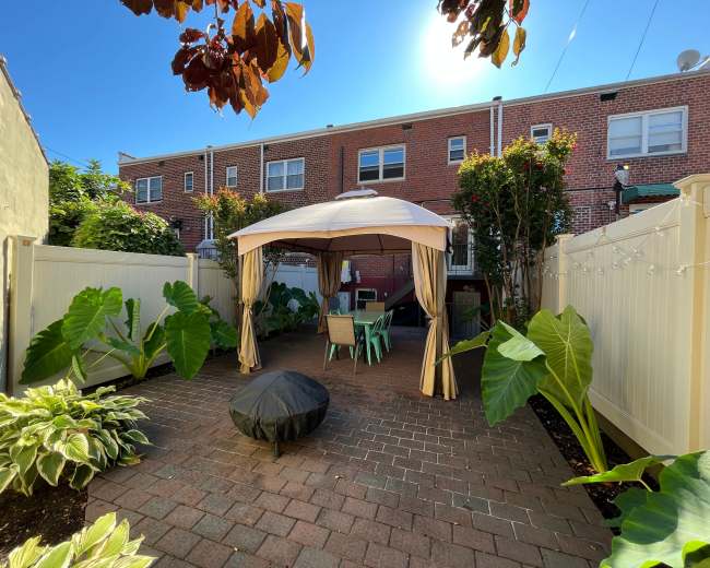 A brick patio features a gazebo surrounded by large foliage and outdoor seating in a residential backyard.