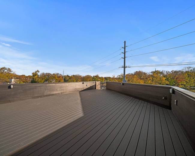 A spacious rooftop deck with wooden flooring overlooks a landscape of trees and power lines under a clear blue sky.