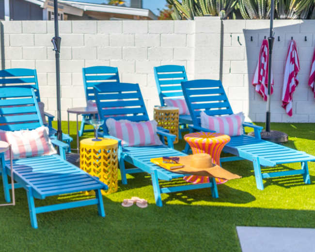 A row of bright blue lounge chairs with colorful side tables and decorative cushions is set up on a grassy patio.