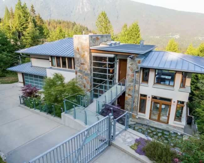 The image shows a modern two-story house with a stone and metal façade, surrounded by trees and mountainous terrain.