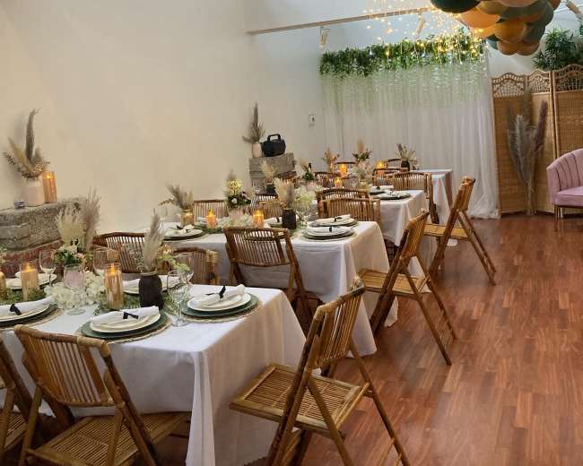 A decorated event space with several tables set for dining, featuring floral centerpieces, candles, and a balloon arrangement hanging above.