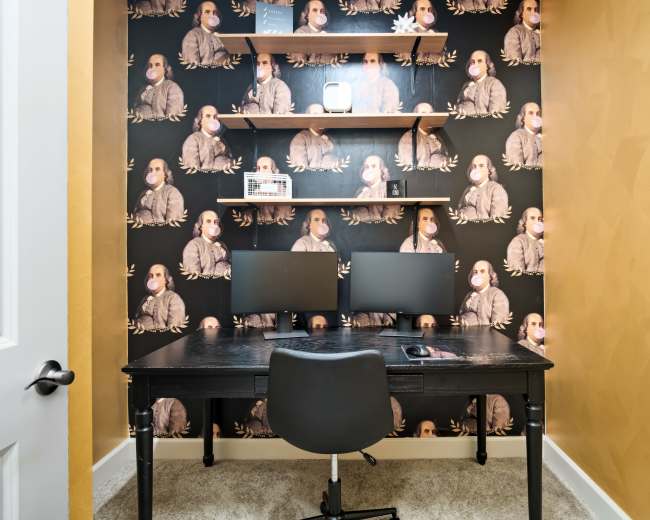 The image shows a small home office featuring a black desk with two computer monitors and a shelf above adorned with patterned wallpaper.