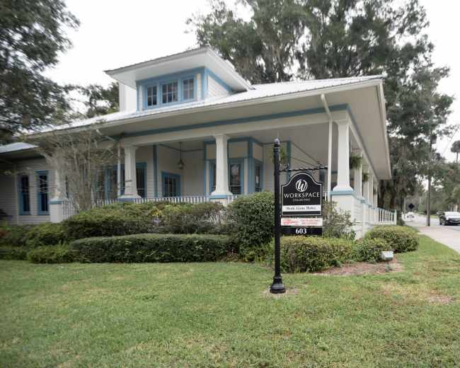 The image shows a large, light-colored building with a porch and a sign that reads "WORKSPACE" in front of a well-manicured lawn.