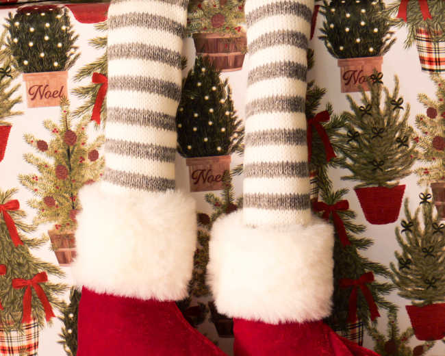A pair of red fuzzy slippers with white cuffs are shown hanging against a festive background of Christmas-themed decorations.