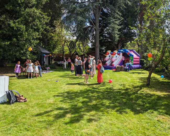 A group of children and adults gather in a grassy yard featuring a bounce house and colorful decorations for a party.