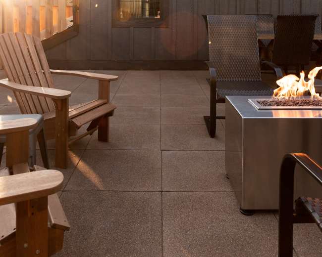 A patio area features wooden adirondack chairs arranged around a fire pit, with a house and fence in the background illuminated by sunlight.