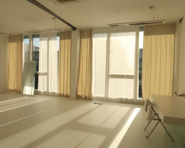 The image shows a bright, spacious room with large windows draped in light curtains, and sunlight casting shadows on the floor.