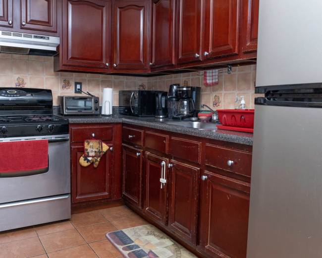 The image shows a kitchen with dark wooden cabinets, a stainless steel stove and refrigerator, and a countertop with various appliances and utensils.