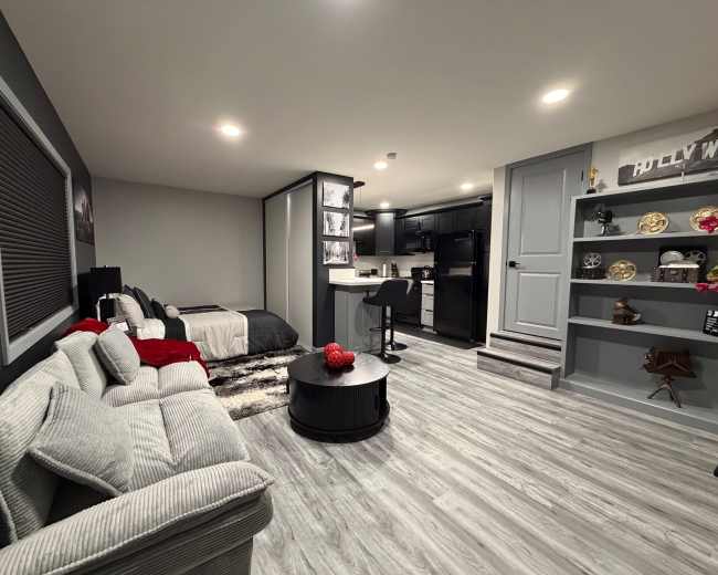 The image shows a modern, minimalistic basement living space with a bed, a cozy couch, and a kitchenette area, accented by decorative shelves.