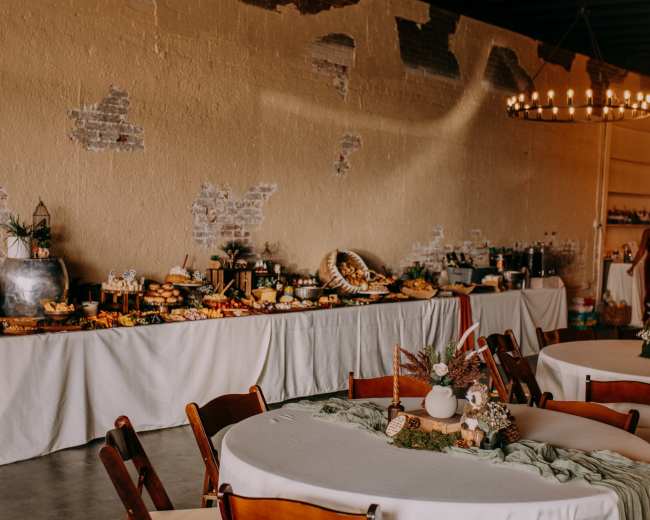A banquet room features round tables with wooden chairs, and a long buffet filled with various dishes and decorative items along a wall.