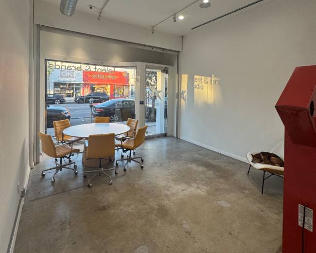 The image depicts a modern, empty office space with a round table and six chairs, large glass doors, and a visible red machine in one corner.