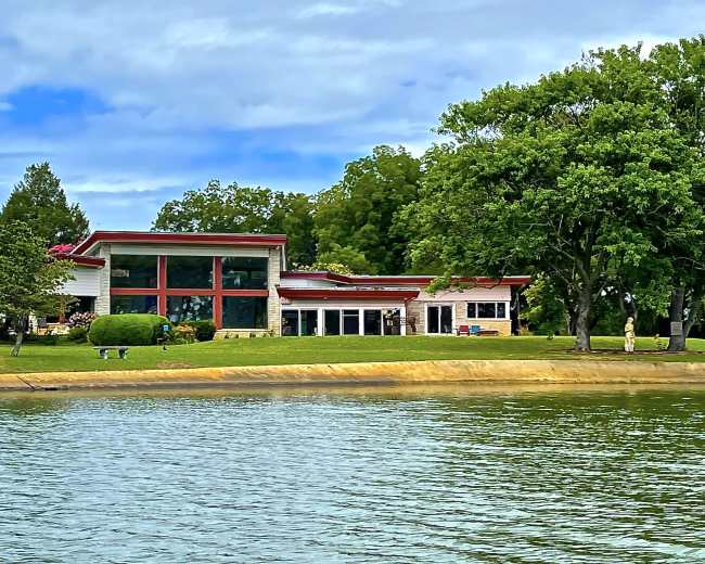 A modern house with large windows and a red roof is situated along the shoreline of a lake, surrounded by trees and a grassy area.