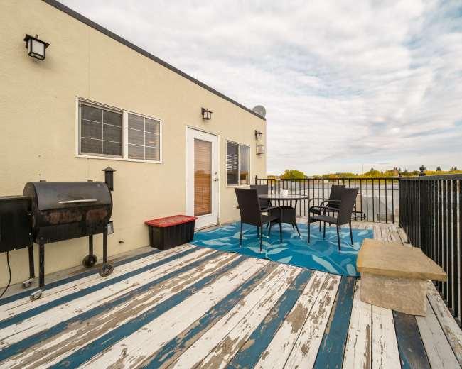 The image shows a rooftop deck featuring a grill, two chairs, and a patterned rug, with a cloudy sky in the background.
