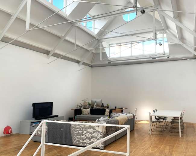 The image shows a spacious, modern room with a high ceiling, featuring a living area with a sofa and television, and a dining table with chairs.