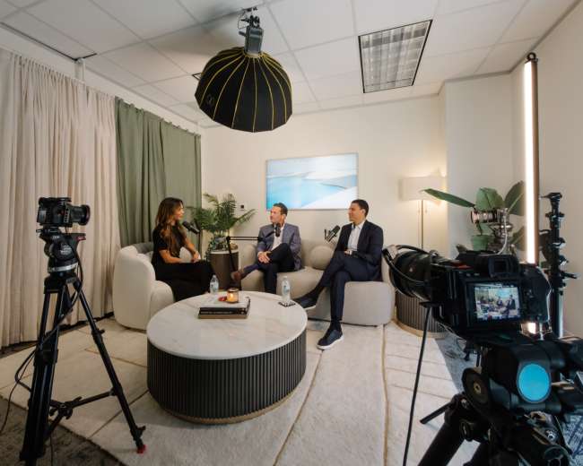 Two men in suits and a woman are seated on a sofa in a well-lit interview setting, surrounded by cameras and a large round table.