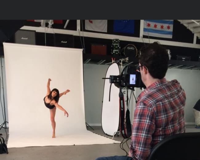 A photographer sits on a chair, capturing a dancer performing an extended pose in front of a plain backdrop in a studio.