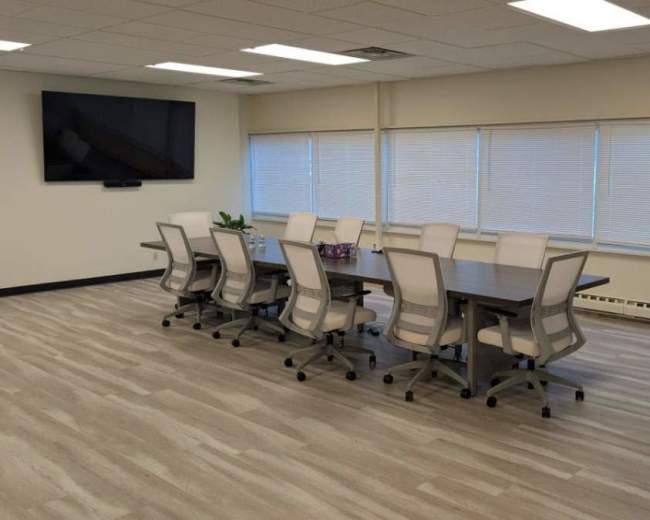 A conference room features a long table surrounded by several light-colored chairs, with windows providing natural light and a large screen mounted on the wall.