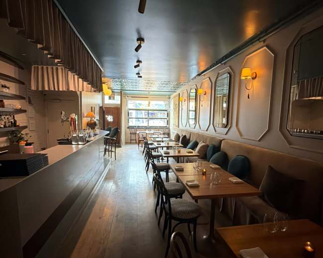 The image shows a long, narrow restaurant interior featuring a bar on one side and tables with chairs arranged along the other side, illuminated by soft lighting.