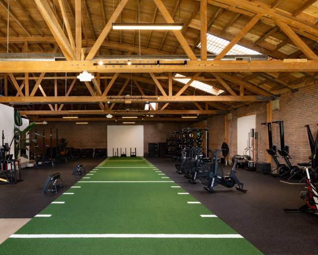 The image shows a spacious gym with a green turf section in the center, surrounded by various fitness equipment on both sides.