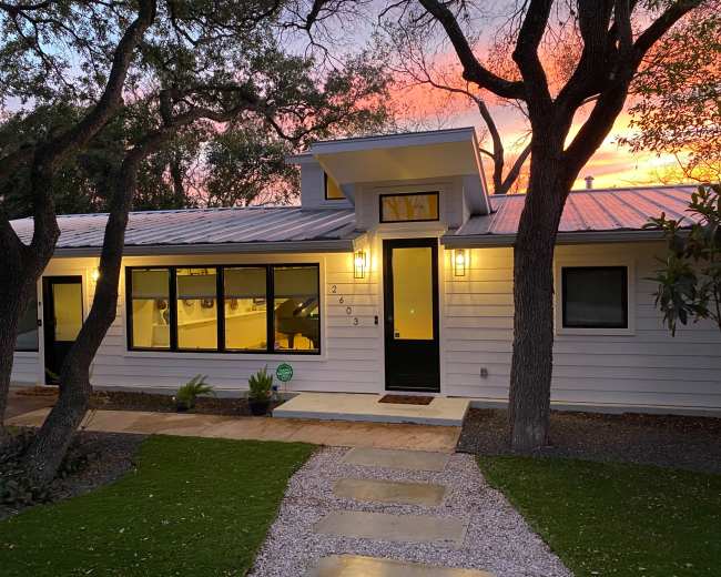 A modern white house with a metal roof and large windows is illuminated at dusk, surrounded by trees and a stone path leading to its entrance.