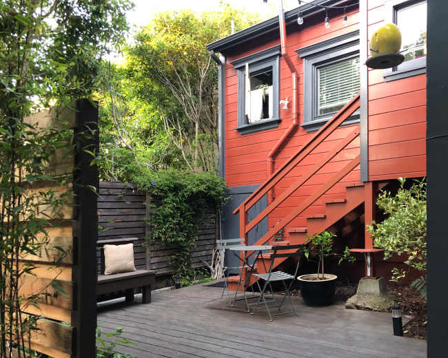 A small outdoor area with a wooden deck, a seating arrangement, and a red house with a staircase and green foliage surrounding it.