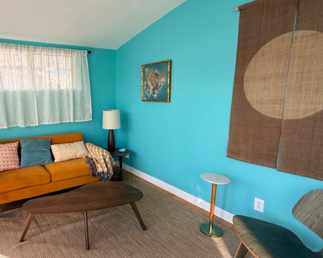 The image shows a brightly colored living room with a teal wall, an orange sofa, a wooden coffee table, and a potted plant.