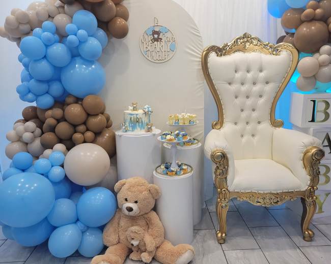 A decorated event space featuring a golden throne chair, a display of dessert treats, and an arrangement of blue and brown balloons.