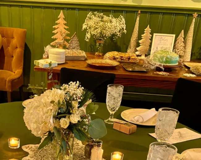 A set dining table is arranged with place settings, a floral centerpiece, and decorations, while a dessert table is adorned with cookies, a cake, and decorative trees in the background.