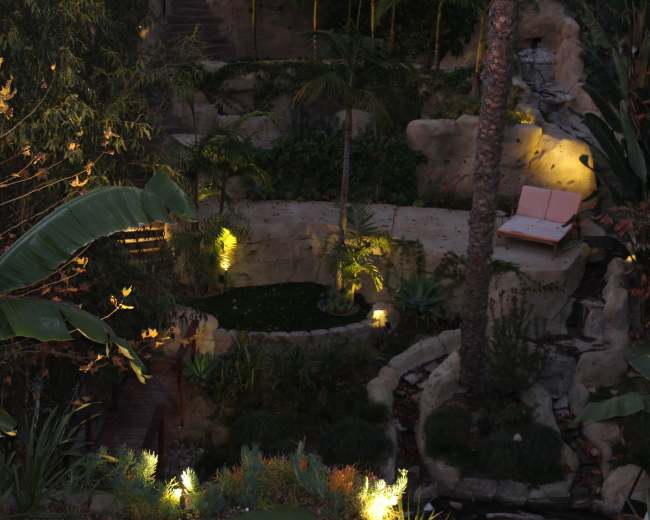 A landscaped garden featuring a stone terrace, a small pond, tropical plants, and a pink lounge chair illuminated by soft lighting.