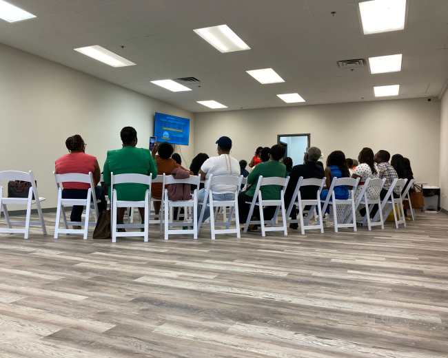 A group of people sit in rows of white chairs facing a screen in a plain room.