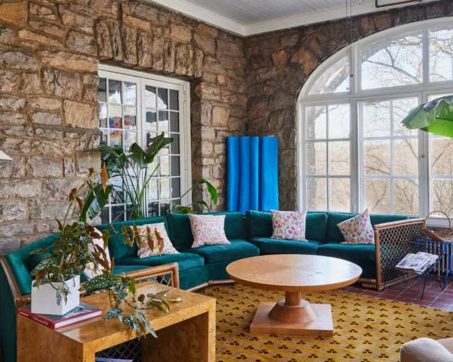 The image shows a brightly lit living room featuring a green sectional sofa, a circular wooden coffee table, and large windows with views of outdoor greenery.