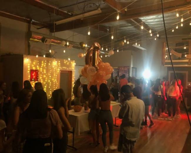 A lively party is taking place in a well-lit venue, featuring balloons, string lights, and groups of people socializing.