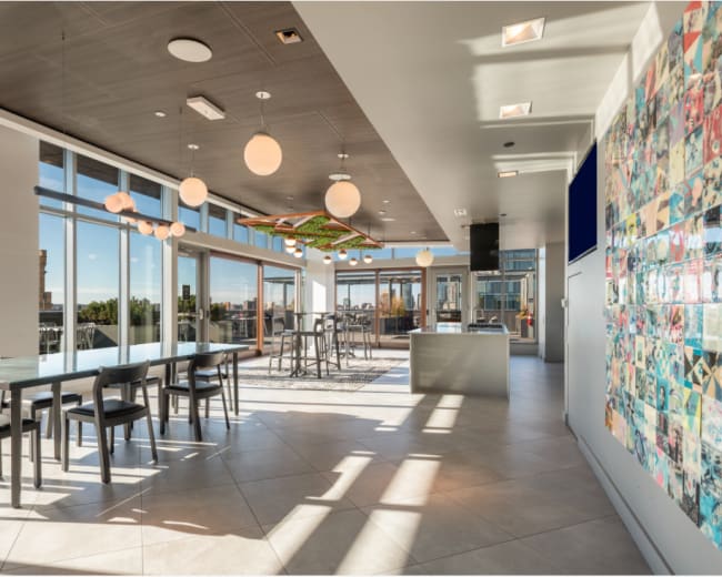 The image depicts a modern open-concept kitchen and dining area featuring large windows, a long table with black chairs, and a colorful wall display.