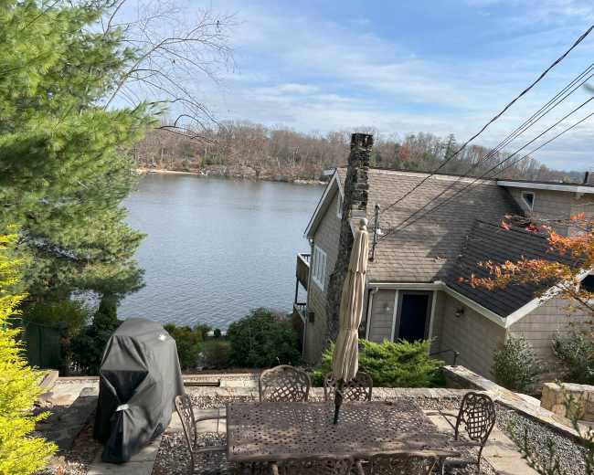 A patio with a dining set and grill overlooks a calm lake surrounded by trees.