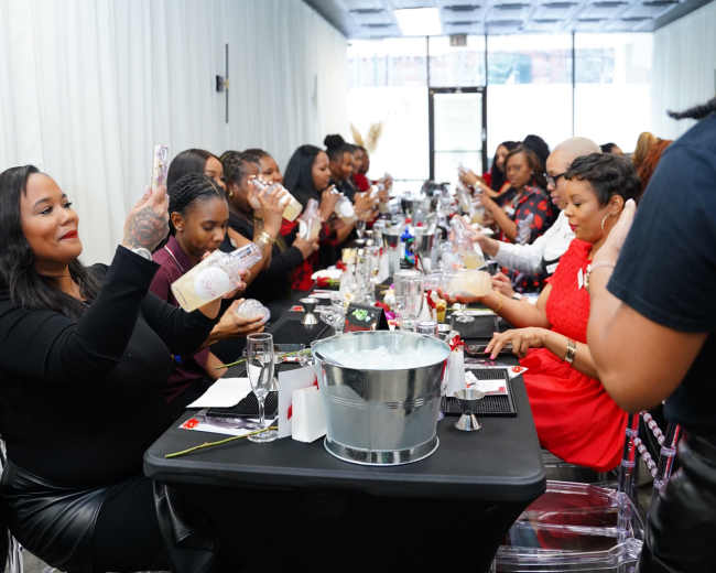 A long table is set for a gathering of people, with attendees raising glasses while seated, in an indoor venue with white drapes and a modern decor.
