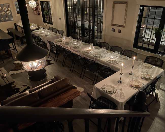 A spacious dining area is set with multiple tables, each covered with white tablecloths and arranged with plates and candles, illuminated by soft lighting.