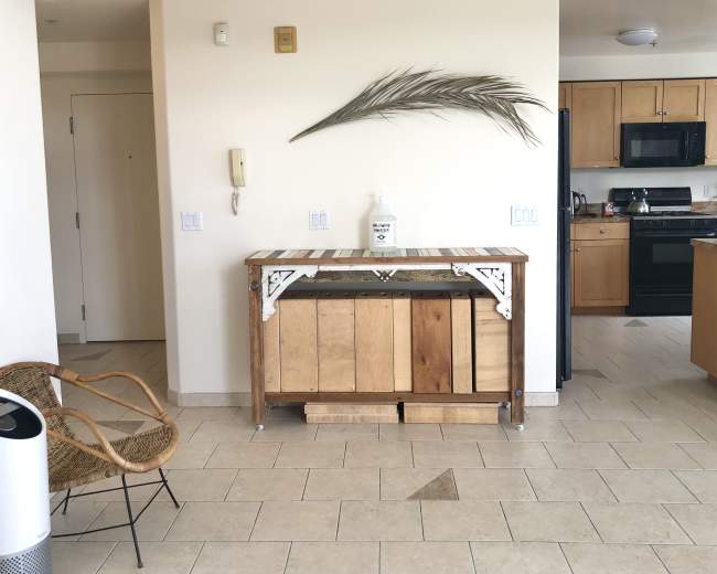 A wooden console table with a decorative palm leaf sits in an open space between a kitchen and a seating area.