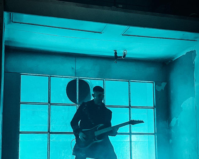 A musician plays an electric guitar in front of a large, illuminated window with a smoky backdrop.