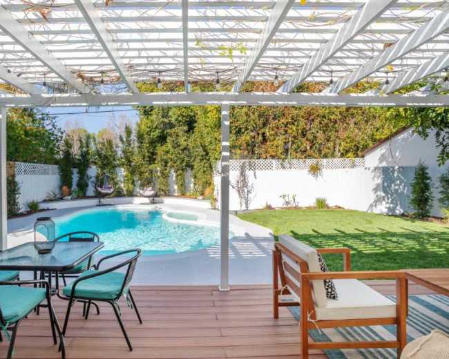 The image shows a backyard with a swimming pool, surrounded by greenery and a shaded seating area with a dining set.