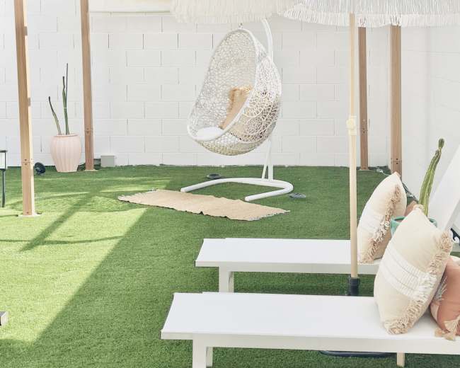 The image shows a backyard space featuring a hanging chair, a large white umbrella, a small rug, and two white benches on artificial grass, surrounded by several potted plants.