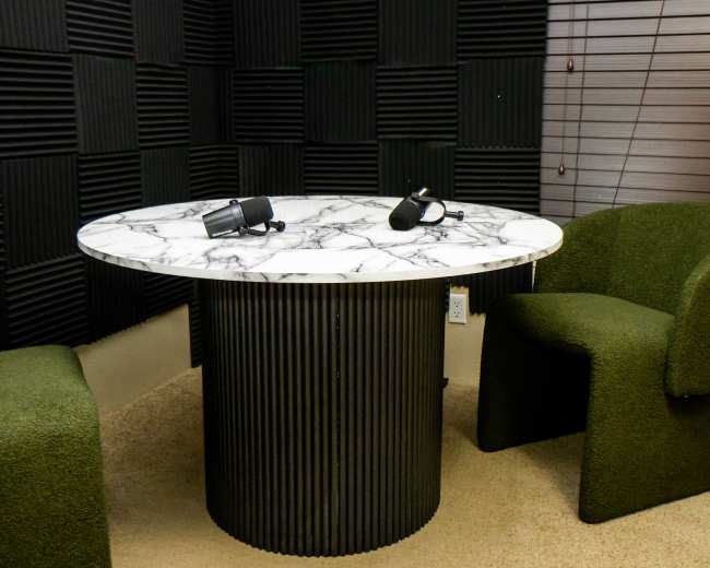 A soundproof room features a round marble table with two green upholstered chairs and microphones positioned on the table.