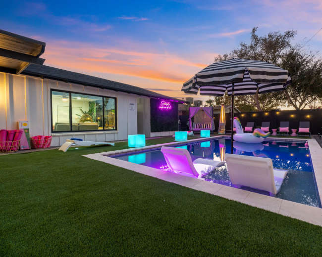 A modern backyard with a swimming pool, surrounded by colorful lounge chairs and an umbrella, set against a sunset sky.