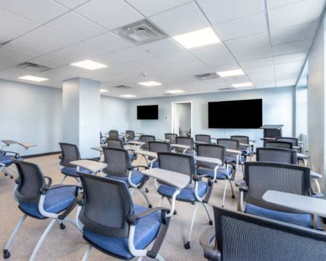 The image shows a modern classroom with rows of movable chairs and desks, two large screens on the walls, and a light blue color scheme.