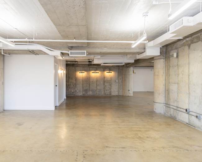 The image shows an empty industrial-style room with exposed concrete walls and a polished concrete floor, illuminated by overhead lighting.