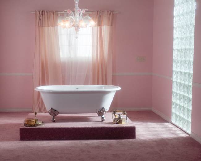 A vintage bathtub sits on a raised platform in a softly lit, pink-walled bathroom adorned with sheer curtains and a chandelier.