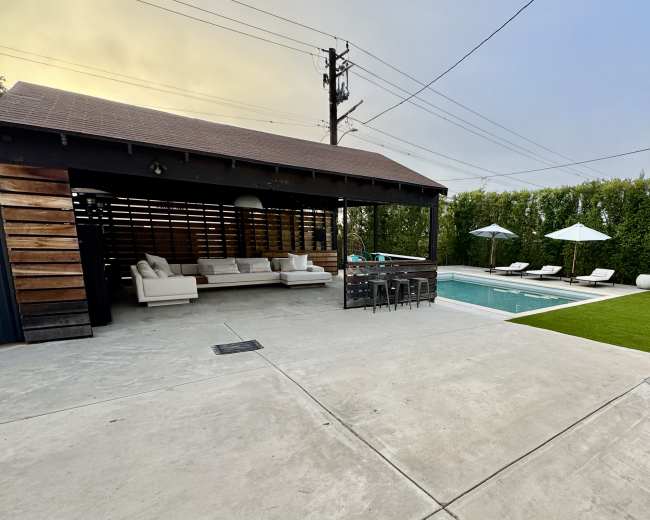 The image shows a modern outdoor area featuring a pool, lounge chairs, a covered seating space, and high hedges providing privacy.