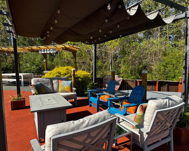 The image shows a patio area with several outdoor seating arrangements, including colorful chairs and a table, under a shaded pergola.