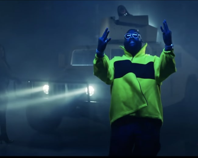A person in a bright yellow jacket stands with arms raised in front of an illuminated vehicle in a foggy setting, while another figure is partially visible in the background.